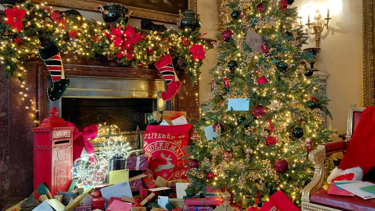 Festively decorated room featuring a large Christmas tree adorned with twinkling lights, red and gold ornaments, and letters to Father Christmas, beside a garland-draped fireplace with stockings, a North Pole post box, sacks of Christmas mail, and piles of wrapped presents.